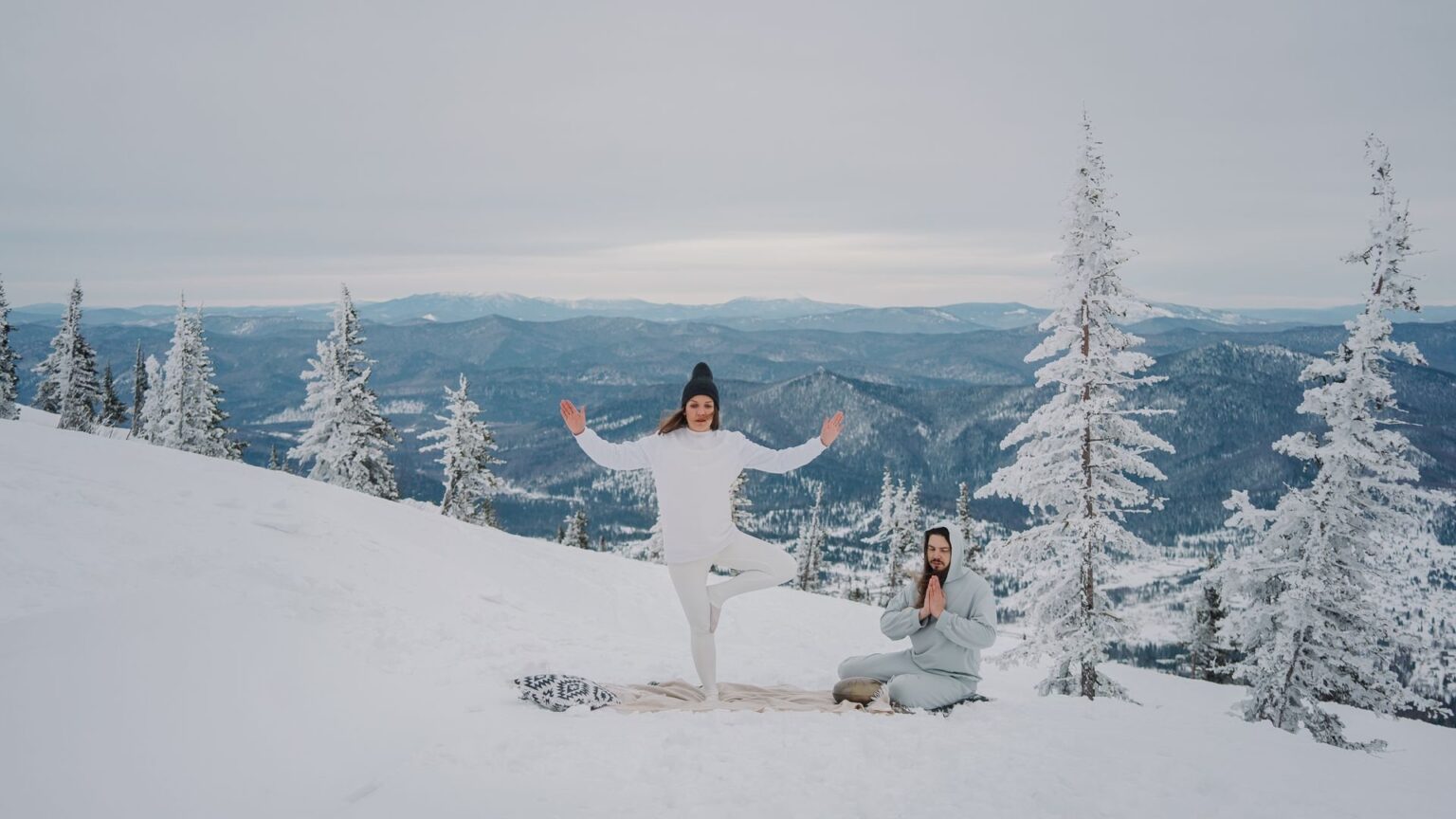 Two people practising yoga and meditation on a snowy mountainside among frosted pines